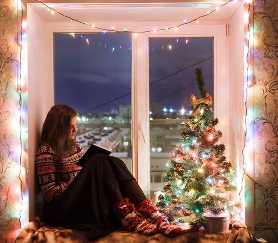 Kvinna sitter och läser en bok i ett julpyntat och nyputsat fönster.